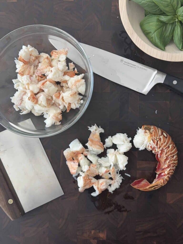 Cooked lobster tail is next to a knife and a glass bowl of already chopped cooked lobster meat. A bowl of basil and a bench scraper are also on the wood counter.