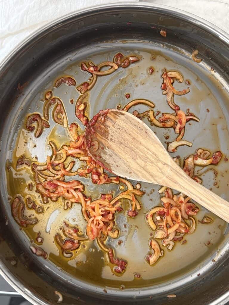 A large skillet with olive oil, sauteed shallot, and tomato paste ready to be stirred with a wooden spoon.