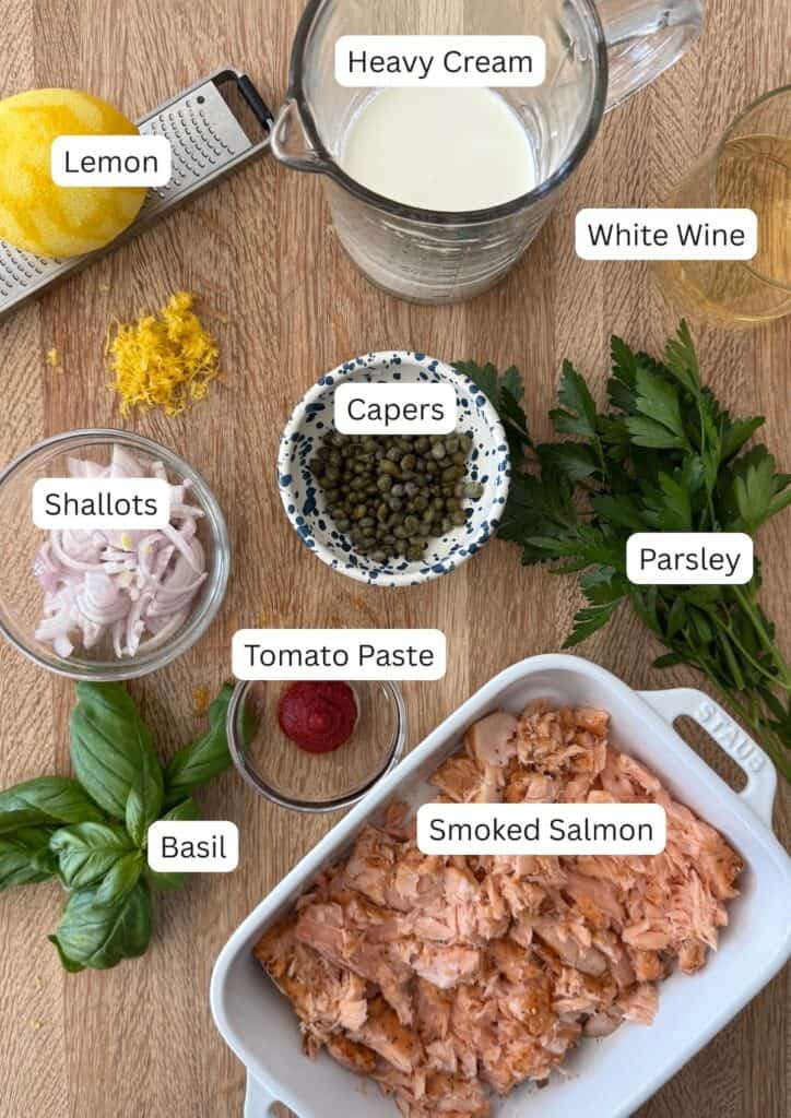 overhead view of ingredients for creamy smoked salmon pasta. the ingredients in the photo are smoked salmon, heavy cream, lemon, white wine, capers, shallot, tomato paste, parsley, and basil.