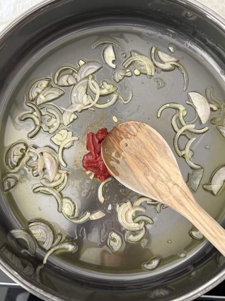 A large skillet with the olive oil, shallot, and tomato paste. a wooden spoon in the skillet to cook the tomato paste.