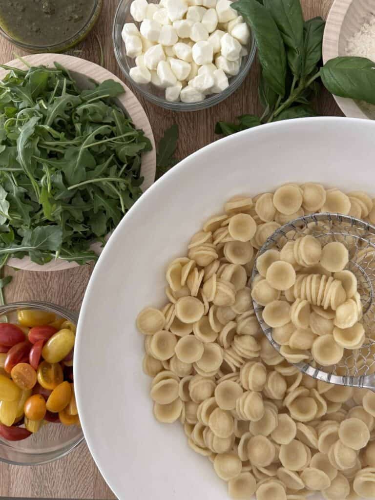Cooked orecchiette pasta is being held by a strainer spoon in a white bowl. Baby arugula, basil, pesto, and cherry tomatoes are in bowls around it.