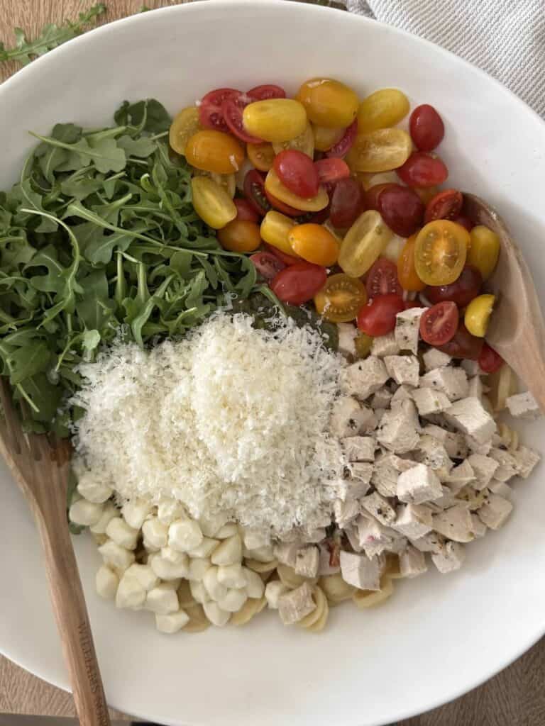 A large white bowl has piles of arugula, cherry tomatoes, diced chicken, parmesan cheese, mozzarella pearls, on top of orecchiette pasta.