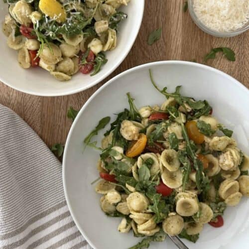 Two bowls of chicken pesto pasta salad. One bowl has a fork and parmesan cheese is in the background.