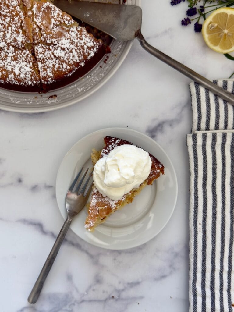 A slice of lemon ricotta cake with whipped cream on a white plate with a fork. The finished lemon ricotta cake is in the background with a cake server.