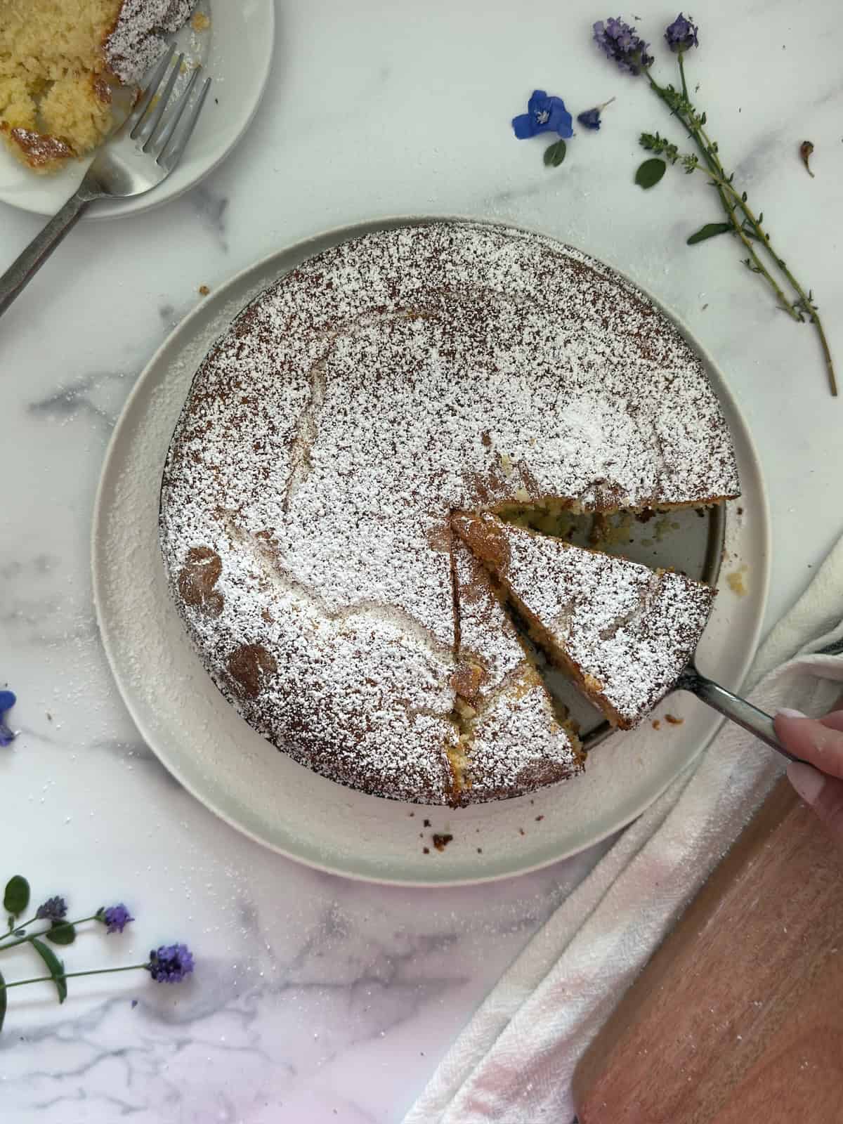 Finished lemon ricotta cake is sliced and on a plate with a cake serve holding one piece. A cut lemon is in the background.