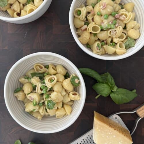 Overhead view of three white bowls filled with pasta e piselli (pasta with peas and pancetta), garnished with fresh basil and Parmesan, set on a dark wooden surface with a block of cheese and grater.