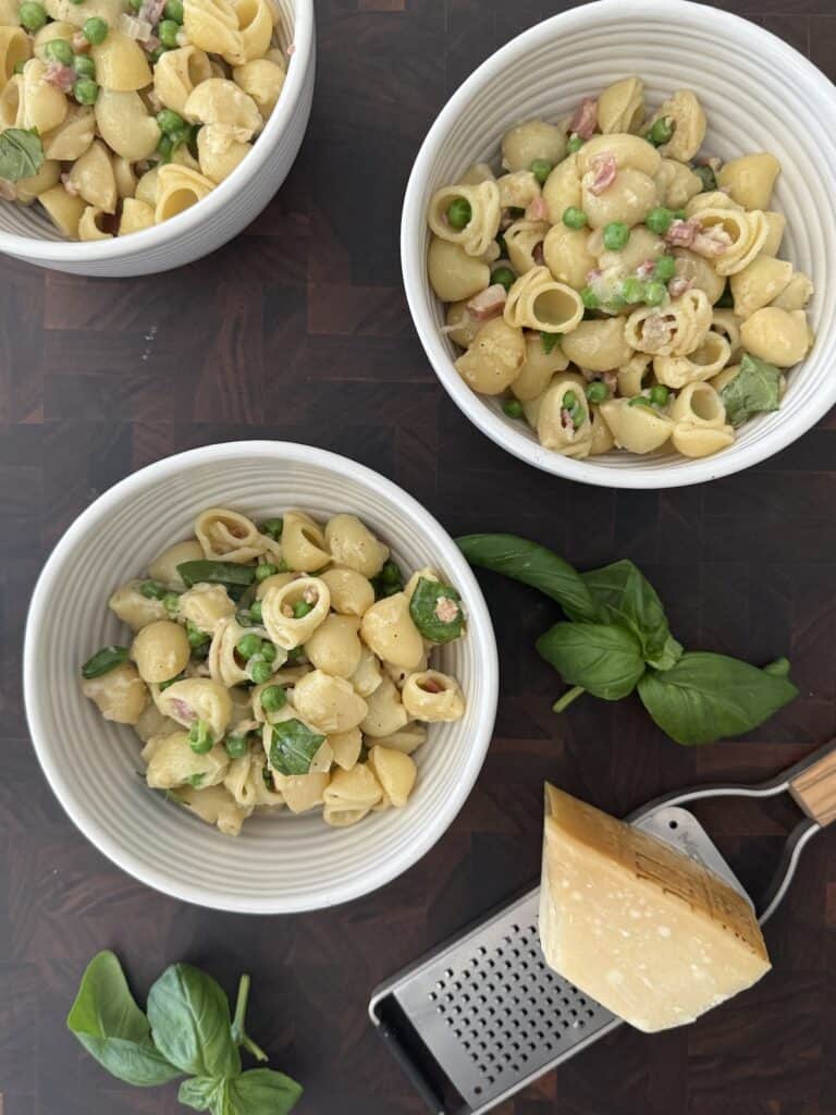 Overhead view of three white bowls filled with pasta e piselli (pasta with peas and pancetta), garnished with fresh basil and Parmesan, set on a dark wooden surface with a block of cheese and grater.