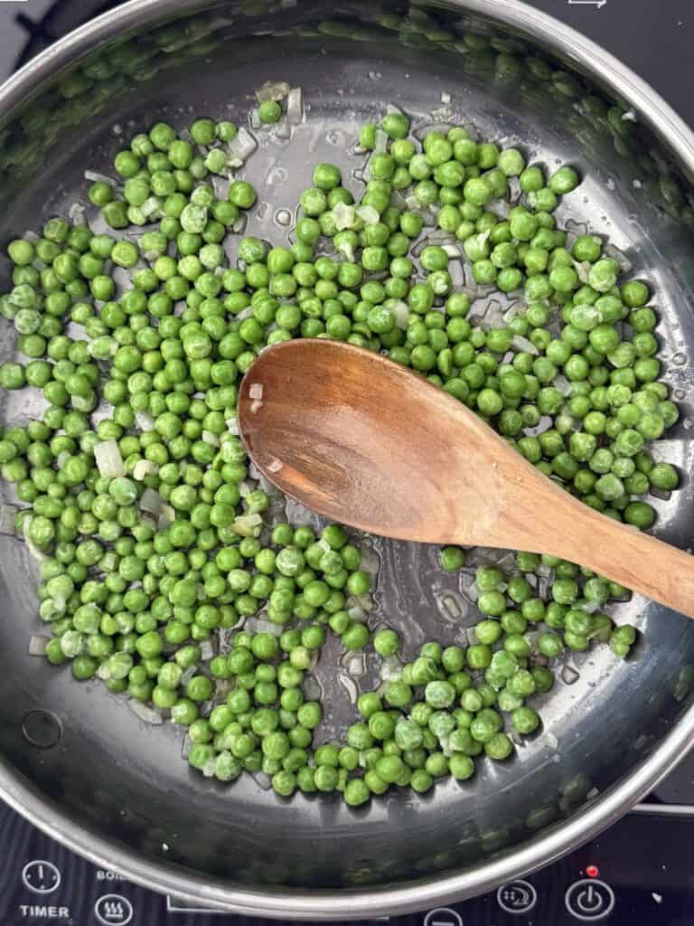 Frozen green peas sautéing in olive oil in a stainless steel skillet, stirred with a wooden spoon as part of pasta e piselli preparation.