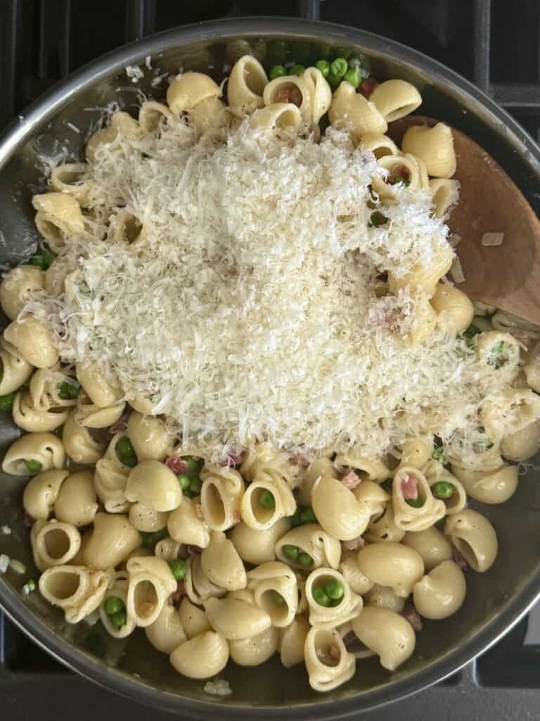 Close-up of pasta e piselli in a skillet, topped with a generous layer of freshly grated Parmesan cheese before mixing.