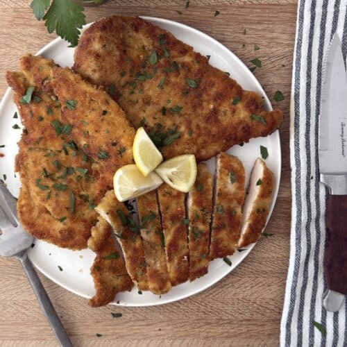 Sliced Italian chicken cutlet on a white plate, garnished with parsley and lemon wedges, served with a striped napkin and fork.