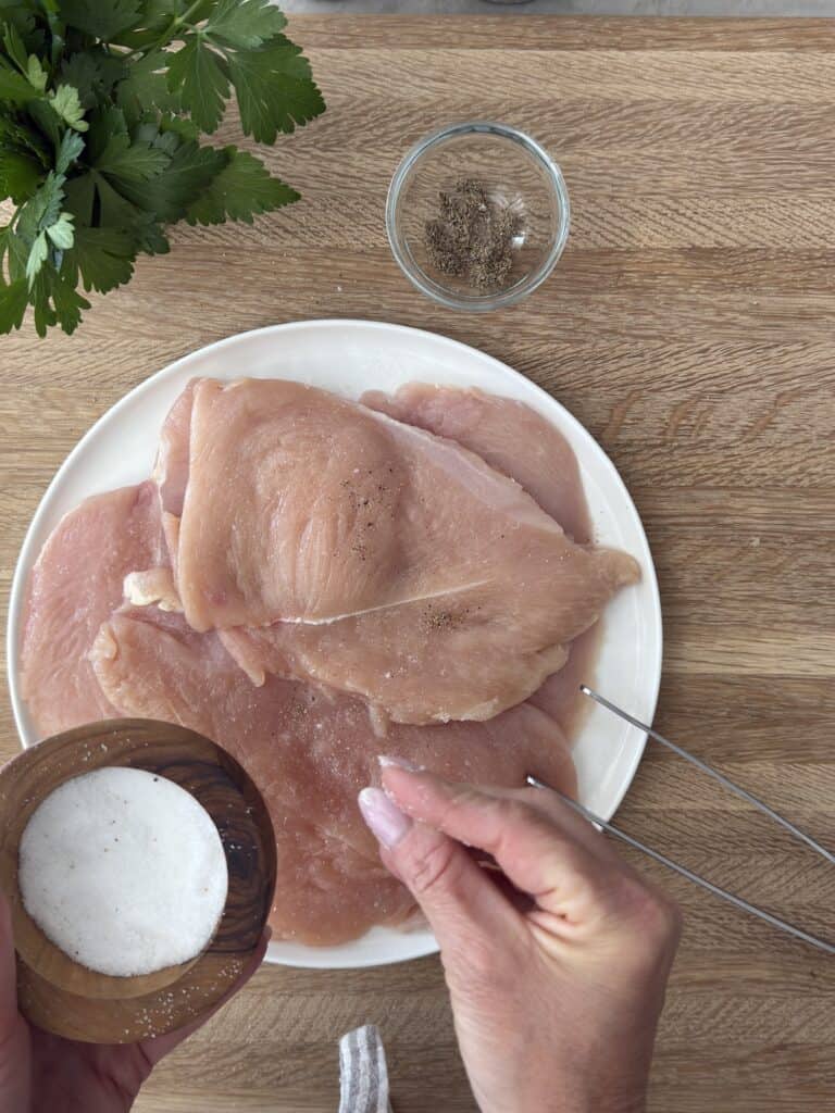 Raw chicken cutlets being seasoned with salt on a wooden board, with black pepper and fresh parsley nearby.