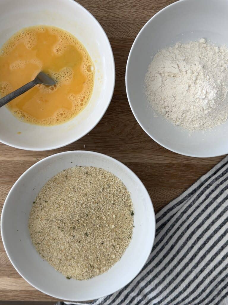 Three bowls with flour, beaten eggs, and breadcrumbs on a wood surface for breading chicken cutlets.