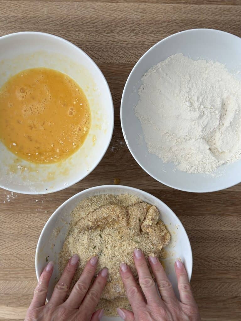 Hands breading chicken cutlets in breadcrumbs, with bowls of beaten eggs, flour, and breadcrumbs on a wooden cutting board.