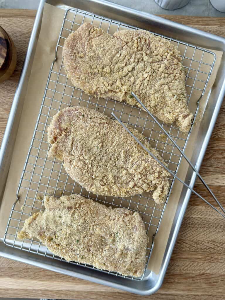 Breaded chicken cutlets arranged on a wire rack over a baking sheet before frying.