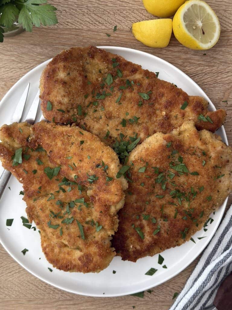 Golden brown Italian chicken cutlets garnished with fresh parsley, served on a white plate with lemon wedges.